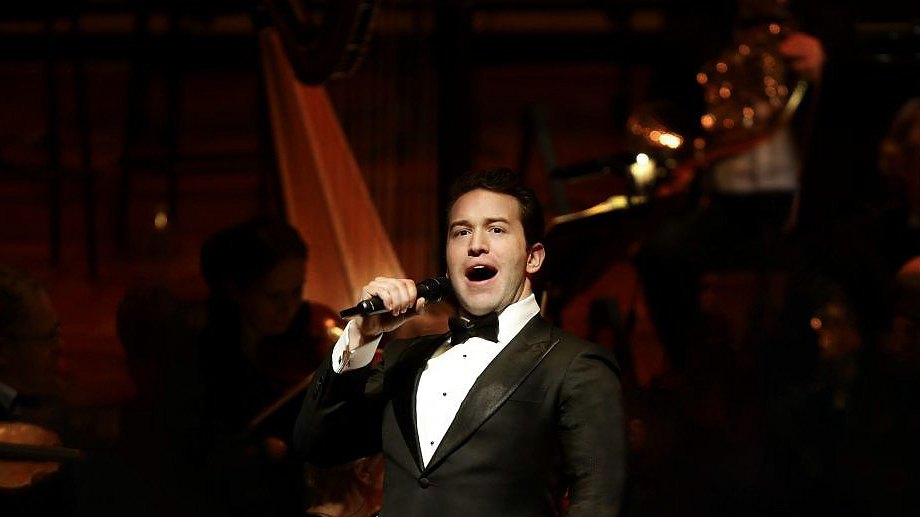 Michael Hewitt singing in a tuxedo with an orchestra behind him